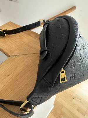 Svart bum bag - Snygg svart bumbag präglat läder med monogrammönster. Väskan har justerbar axelrem och guldfärgade metalldetaljer, inklusive dragkedja och spännen. 