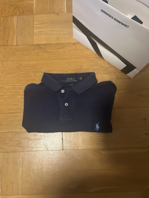 Mörkblå pikétröja från Polo Ralph Lauren - Snygg mörkblå pikétröja från Polo Ralph Lauren med klassisk krage och två knappar framtill. Tröjan har korta ärmar och den ikoniska logon broderad i ljusblått på bröstet. Perfekt för en avslappnad och stilren look.
