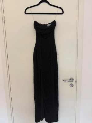 Svart glittrig långklänning med slits - Supersnygg svart långklänning med glittrigt tyg. Klänningen har en hög slits på ena sidan och en smickrande, åtsittande passform. Perfekt för dig som vill sticka ut på bal eller fest! Materialet känns mjukt och följsamt. Storleken är UK 4, passar dig som har XS, men kan möjligtvis passa dig som har S. Materialet är strechigt.