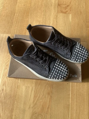 Christian Louboutin svarta sneakers - Svarta sneakers från Christian Louboutin med coola silverfärgade nitar på tån. Skorna har en klassisk låg siluett, snörning och vit sula. Materialet är troligtvis mocka eller sammet, vilket ger en lyxig känsla. Perfekta för dig som vill sticka ut med din stil.