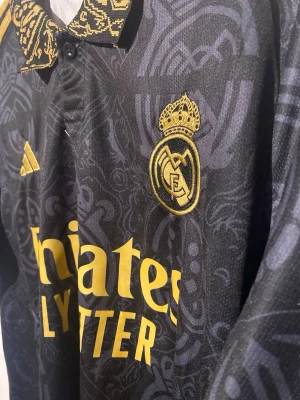 Real Madrid svart matchtröja Adidas - Snygg svart Real Madrid matchtröja från Adidas med guldfärgade detaljer och klubbmärke på bröstet. Tröjan har korta ärmar, mönstrad krage och ärmslut samt Adidas-logga och sponsortryck i gult. Tillverkad i lätt och ventilerande material. Storlek S (denna finns inne hos oss nu)