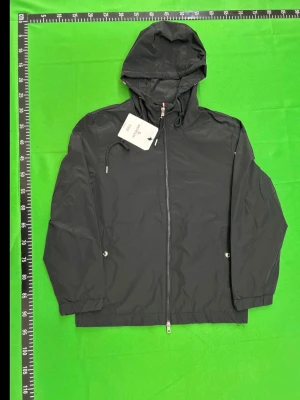 Svart vindjacka från Moncler - Snygg svart vindjacka från Moncler med huva och dragkedja framtill. Jackan har två sidofickor med tryckknappar, justerbar dragsko i huvan och Moncler-logga på ärmen. Materialet är tunt och vattenavvisande, perfekt för blåsiga dagar.