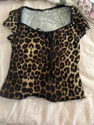 Leopardmönstrad blus med rosett - Snygg blus med leopardmönster och transparent mesh upptill. Dekorativ svart rosett vid halsringningen och korta ärmar. Perfekt för dig som vill sticka ut med ett djurmönster. Luftigt material och lätt figurnära passform.