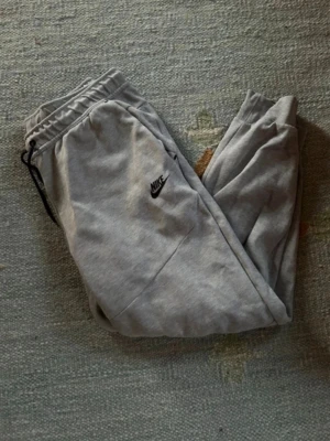 Nike tech fleece byxor - Säljer ett par gråa mjukisbyxor från Nike med svart logga på benet. Byxorna har resår i midjan med dragsko, muddar vid bensluten och en dragkedjeförsedd ficka på sidan. Perfekta för chill eller träning.
