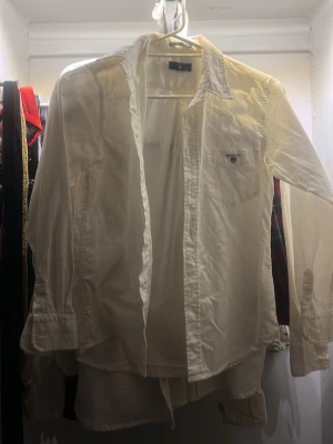 Vit skjorta från Gant med broderad logga, skjorta för barn ! - Klassisk vit skjorta från Gant med lång ärm och bröstficka. Skjortan har broderad Gant-logga och sköld på fickan, knappar framtill och en tidlös krage. Perfekt att styla lager på lager eller bära som den är.