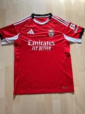 Benfica röd matchtröja Adidas - Röd Benfica matchtröja från Adidas med vita detaljer och svarta inslag vid halsen. Tröjan har klubbens emblem, tre stjärnor ovanför, och sponsorloggan 'Emirates Fly Better' på bröstet. Korta ärmar med vita och svarta inslag samt Adidas-logga på axeln.