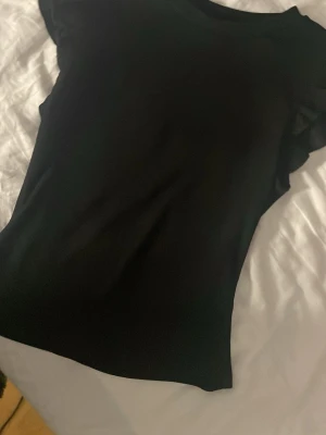 Svart topp med volangärmar - Snygg svart topp med korta volangärmar och rund hals. Tunn och mjuk känsla! Enkel men ändå trendig design som passar till många olika looks. 🤩