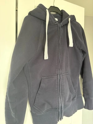 Mörkblå zip hoodie med vita snören och byxor/russedress - Mysig mörkgrå hoodie med dragkedja och stora fickor framtill. Har huva med vita, breda dragsnören och ribbade muddar vid ärmslut och nederkant. Perfekt för en avslappnad och chill stil. Materialet känns mjukt och bekvämt, troligen bomullsblandning.