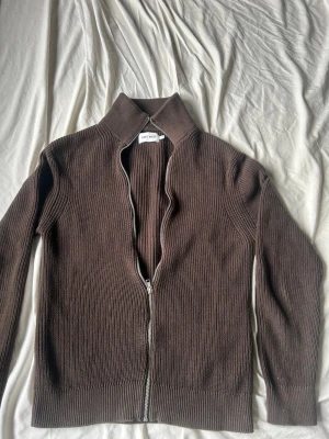 Brun ribbstickad ziptröja East West - Brun ribbstickad tröja från East West med dragkedja framtill och hög krage. Tröjan har långa ärmar och en clean, minimalistisk look som passar perfekt till lager på lager. Materialet är tjockt och mjukt för extra komfort. Storlek s men passar även xs
