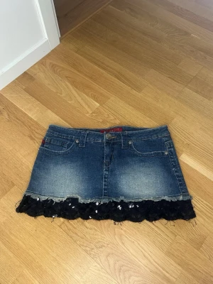 Jeanskjol med spets och paljetter - Säljer en snygg blå jeanskjol från YMI med svart spets och paljetter längst ner. Kjolen har klassiska jeansfickor fram och bak, bälteshällor och knappstängning. Perfekt för dig som vill ha en unik twist på din denimstil!