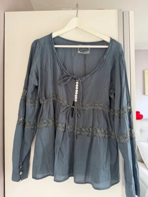 blus tunika  med spetsdetaljer - Supersöt blå bohemisk blus från Fira med långa ärmar, knytband fram och dekorativa vita knappar. Blusen har spetsdetaljer över bröstet och ärmarna samt lätt rynkad midja för en avslappnad look. Perfekt till jeans eller kjol.