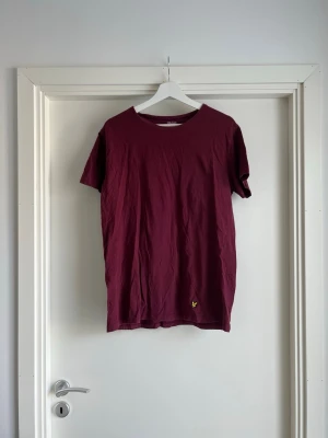Vinröd t-shirt från Lyle & Scott - Säljer en vinröd burgundy röd t-shirt från Lyle & Scott med klassisk rund hals och korta ärmar. T-shirten har en liten gul logga broderad längst ner på framsidan. Tillverkad i mjuk bomull och har en clean, enkel look. Storlek L / Large. Måtten finns. 