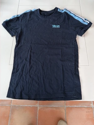 Marinblå t-shirt med ljusblå ränder - Snygg marinblå t-shirt från 1910 med ljusblå ränder längs axlarna. Klassisk rund hals och korta ärmar. Perfekt för en avslappnad stil och enkel att matcha med jeans eller shorts.