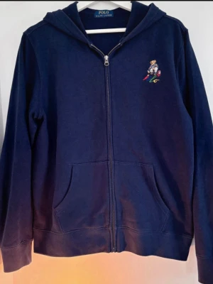 Marinblå hoodie från Polo Ralph Lauren - Mörkblå hoodie med dragkedja från Polo Ralph Lauren. Klassisk björnlogga broderad på bröstet och två fickor framtill. Tillverkad i mjukt bomullsmaterial och har en skön huva. Perfekt för en avslappnad och stilren look.