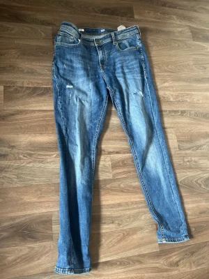 Ett par fräscha Jack & Jones Jeans med slitningar - Säljer ett par blå skinny jeans med slitningar på låren och knäna. Jeansen har klassisk femficksmodell, normalhög midja och dragkedja med knapp. Materialet är denim i bomull med lite stretch för skön passform.