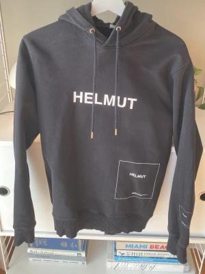 Svart hoodie från Helmut - Svart hoodie från Helmut med vit text och grafiska detaljer på bröstet, magen och ena ärmen. Klassisk huva med dragsko och ribbade muddar vid ärmslut och nederkant. Tillverkad i mjukt bomullsmaterial för en avslappnad och trendig look.
