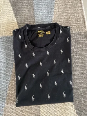 Svart t-shirt från Polo Ralph Lauren - Svart t-shirt från Polo Ralph Lauren med små vita broderade loggor över hela tröjan. Klassisk rund halsringning och korta ärmar. Tillverkad i mjuk bomull som känns skön mot huden. Perfekt för en clean och stilren look.