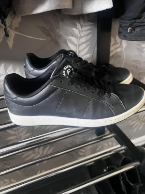 Svarta sneakers från Le Marune - Stilrena svarta sneakers från Le Marune med lågt skaft och klassisk vit sula. Skorna har svarta skosnören och är tillverkade i slätt konstläder. Perfekta för en clean och enkel look.