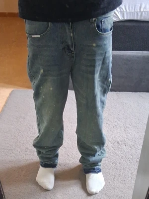 Blå raka jeans med vintagekänsla - Säljer ett par blå jeans med rak passform och klassisk femficksdesign. Jeansen har en avslappnad look med slitningar och färgstänk för en cool vintage vibe. Perfekta för dig som gillar streetwear och en avslappnad stil.