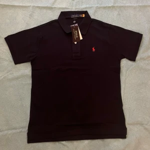Svart pikétröja från Polo Ralph Lauren - Klassisk svart pikétröja från Polo Ralph Lauren med korta ärmar och röd broderad logga på bröstet. Tröjan har krage och knappar framtill, perfekt för en clean och stilren look.