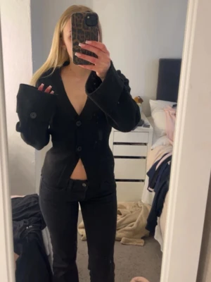 Svart stickad kofta med vida ärmar - Snygg svart stickad kofta med djup V-ringning och stora, vida ärmar med dekorativa knappar. Figurnära passform och tre knappar framtill. Perfekt för en trendig och edgy look. Materialet ser ut att vara en mjuk och något stretchig textil.