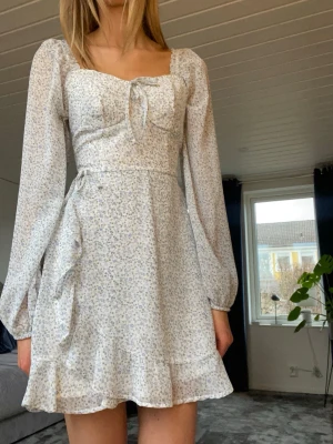 Omlott klänning - Söt kort omlottklänning i vitt med små blå och lila blommor från hollister. Klänningen har långa ärmar, volang nertill och knytning i midjan. Endast använd vid ett tillfälle.