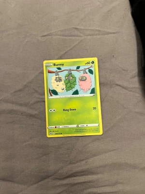 Pokémonkort Burmy - Säljer ett Pokémonkort med Burmy-motivet. Kortet har en grön ram och visar tre olika Burmy i olika färger hängande från en gren. Kortet är gjort av papper/kartong och har attacken Hang Down. Perfekt för samlare eller Pokémonfans.