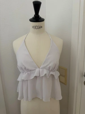 Vit volang topp med knyt i nacken - Supersöt vit topp i XS med volang och djup v-ringning. Toppen har knyt i nacken och är ärmlös med öppen rygg. Tillverkad i ett lätt och luftigt material som ger en flowy känsla. Skickas eller hämtas på Hägerstensåsen. 🌸