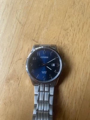 Citizen klocka med blå urtavla - Stilren Citizen klocka med blå urtavla och datumvisning. Armbandet är i silverfärgat metall och klockan har tydliga siffror samt klassisk rund form. Perfekt accessoar för dig som gillar enkel och tidlös design. Extra länkar till armbandet medföljer.