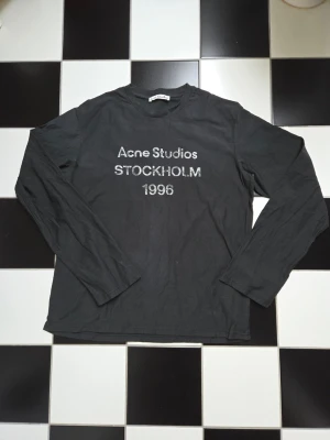 Svart långärmad topp Acne Studios - Svart långärmad topp från Acne Studios med tryck 'Acne Studios STOCKHOLM 1996' på bröstet. Tillverkad i mjuk bomull med normal passform och rund hals. Perfekt för en clean och stilren look.