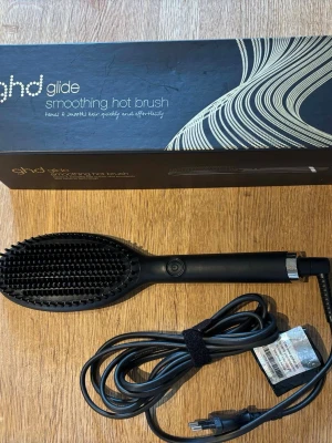 ghd Glide smoothing hot brush borste  - En svart elektrisk värmeborste från ghd, modell Glide smoothing hot brush. Borsten har täta piggar och en lång sladd med kardborreband för enkel förvaring. Perfekt för att snabbt och smidigt styla och släta ut håret. Levereras med originalkartong.