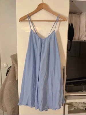 Ljusblå sommarklänning med smala band - Säljer en ljusblå, luftig klänning med tunna axelband och v-ringning. Klänningen har en avslappnad passform och är tillverkad i ett lätt tyg som ger en skön känsla. Perfekt för varma dagar och enkel att styla med sneakers eller sandaler.