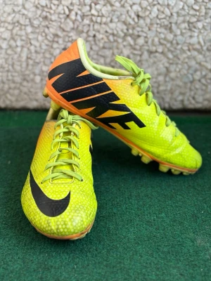 Nike Mercurial fotbollsskor gul/orange - Nike Mercurial fotbollsskor i en grymt snygg gul och orange färg med stora svarta loggor. Skorna har snörning och är tillverkade i syntetmaterial med mönstrad yta för extra grepp. Perfekta för dig som vill sticka ut på planen!