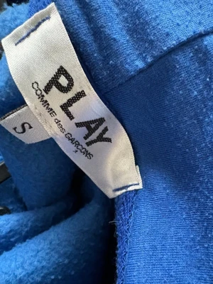 Blå hoodie från Comme des Garçons - Blå hoodie med dragkedja från Comme des Garçons Play. Har ett svart hjärta med ögon broderat på bröstet, ribbade muddar och fickor framtill. Tillverkad i mjukt material och har huva med snörhål.