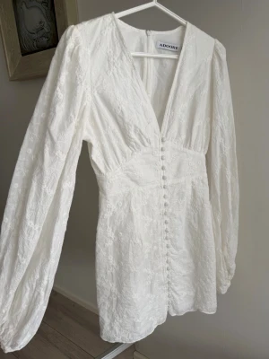 Vit spetsklänning från Adoore - Supersnygg vit kortklänning från Adoore med v-ringning, långa ärmar och dekorativa knappar framtill. Klänningen har fina spetsdetaljer och markerad midja som ger en feminin siluett. Perfekt för sommarens alla tillfällen!