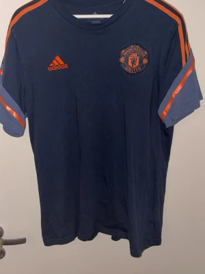 Manchester United t-shirt från Adidas - Mörkblå Manchester United t-shirt från Adidas med orange detaljer och logga på bröstet. Klassiska Adidas-ränder i orange längs axlarna och ärmarna. Perfekt för dig som älskar fotboll och vill ha en snygg supportertröja. Tillverkad i mjukt material.