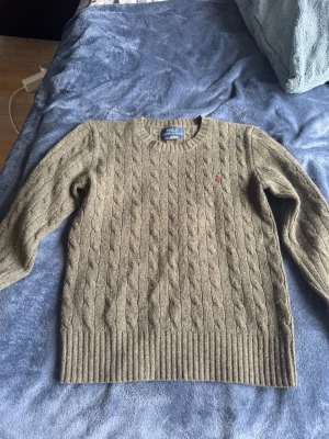 Kabelstickad tröja från Polo Ralph Lauren - Kabelstickad tröja i olivgrönt från Polo Ralph Lauren. Klassisk rund halsringning, ribbade muddar och nederkant samt den ikoniska lilla röda logotypen broderad på bröstet. Tillverkad i mjuk ullblandning som håller dig varm och snygg. Tröjan är i storlek 10- 12 år. Ca 140. Hör av dig vid minsta fundering. 