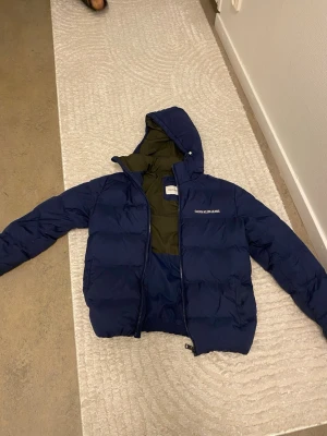 Blå pufferjacka från Calvin Klein - Snygg blå pufferjacka från Calvin Klein med huva och dragkedja. Jackan har en olivgrön insida och diskret logga på bröstet. Perfekt för kalla dagar och har en modern, boxig passform. Materialet är syntet och jackan är vadderad för extra värme.