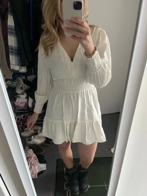 Vit kort klänning från Zara - Supersöt vit kort klänning från Zara med v-ringning, långa ärmar och smock i midjan. Klänningen har volangkant nertill och är gjord i ett luftigt linnematerial. Perfekt för varma dagar och ger en fräsch och trendig look.