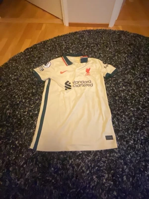Liverpool bortatröja Nike beige - Snygg beige Liverpool FC bortatröja från Nike med gröna detaljer och röda inslag på kragen. Tryck med 'VIRGIL' och nummer 4 på ryggen. Premier League-märke på ärmen och sponsorlogga framtill. Perfekt för dig som älskar fotboll och vill stötta ditt lag.