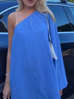 Blå oneshoulder klänning - Snygg blå oneshoulder klänning med lös passform och lång ärm på ena sidan. Perfekt för dig som vill sticka ut med en enkel men trendig look. Materialet känns lätt och luftigt, vilket gör den skön att bära under varma dagar. Köpt på Plick den hade två mindre rosa fläckar på ryggen 