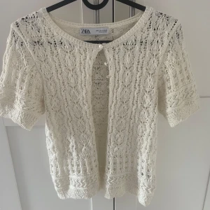 Vit stickad kofta från Zara med pärlknappar - Superfin vit stickad kofta från Zara med korta ärmar och pärlknappar framtill. Blusen har ett detaljerat spetsmönster och rund halsringning. Endast använd 2 gånger 
