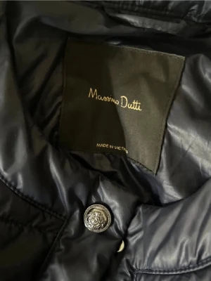 Mörkblå jacka Massimo Dutti XS - Stilren mörkblå vindjacka från Massimo Dutti i glansigt material. Jackan har knappdetaljer med logotyp och klassisk krage. Perfekt för dig som vill ha en lätt och snygg jacka till vardagslooken.