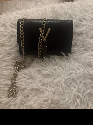 Svart handväska från Valentino - Snygg svart handväska från Valentino med guldfärgad kedja och stort V i metall framtill. Väskan är i läder med struktur och har dekorativ tofsdetalj. Perfekt accessoar för att lyfta din outfit.