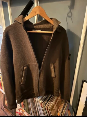 Brun hoodie/cardigan i ull från Stone Island - Säljer en brun hoodie/cardigan i ull från Stone Island med dragkedja framtill och två fickor. Klassisk logotyp på ärmen och ribbade muddar. Perfekt för dig som gillar streetwear och vill ha något både snyggt och bekvämt.