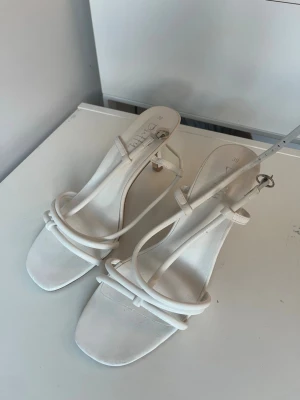 Klackar  - Säljer ett par vita klackar. Skorna har en elegant smal klack och justerbart spänne vid ankeln. Perfekta för avslutning eller fest.