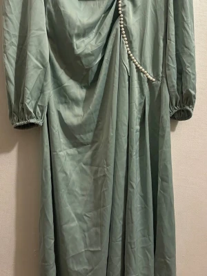 Ljusgrön långklänning med pärldetalj - Elegant ljusgrön långklänning med puffiga långärmar och vacker drapering framtill. Klänningen har en dekorativ rad med pärlor längs ena sidan och resår vid ärmsluten. Perfekt för dig som vill sticka ut med en unik detalj.