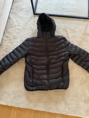 Svart pufferjacka med huva - Säljer en svart värme jacka med avtagbar huva. Jackan har två sidofickor med dragkedja och quiltad design. Perfekt för kalla dagar och riktigt skön att bära. 