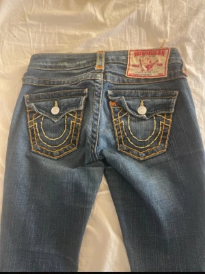 True Religion jeans - Säljer ett par blå jeans från True Religion med ikoniska kontrastsömmar och dekorativa ficklock bak. Klassisk femficksmodell med raka ben och slitna detaljer. Jeansen har en patch med logga bak i midjan och silverfärgade knappar. Jeansen är köpta från Vinted, men är tyvärr för små för mig😇