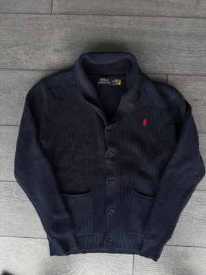 Mörkblå kofta från Polo Ralph Lauren - Mörkblå stickad kofta från Polo Ralph Lauren med knäppning framtill och två fickor. Klassisk design med ribbstickade muddar och röd broderad logga på bröstet. Perfekt för lager-på-lager och snygg till jeans.
